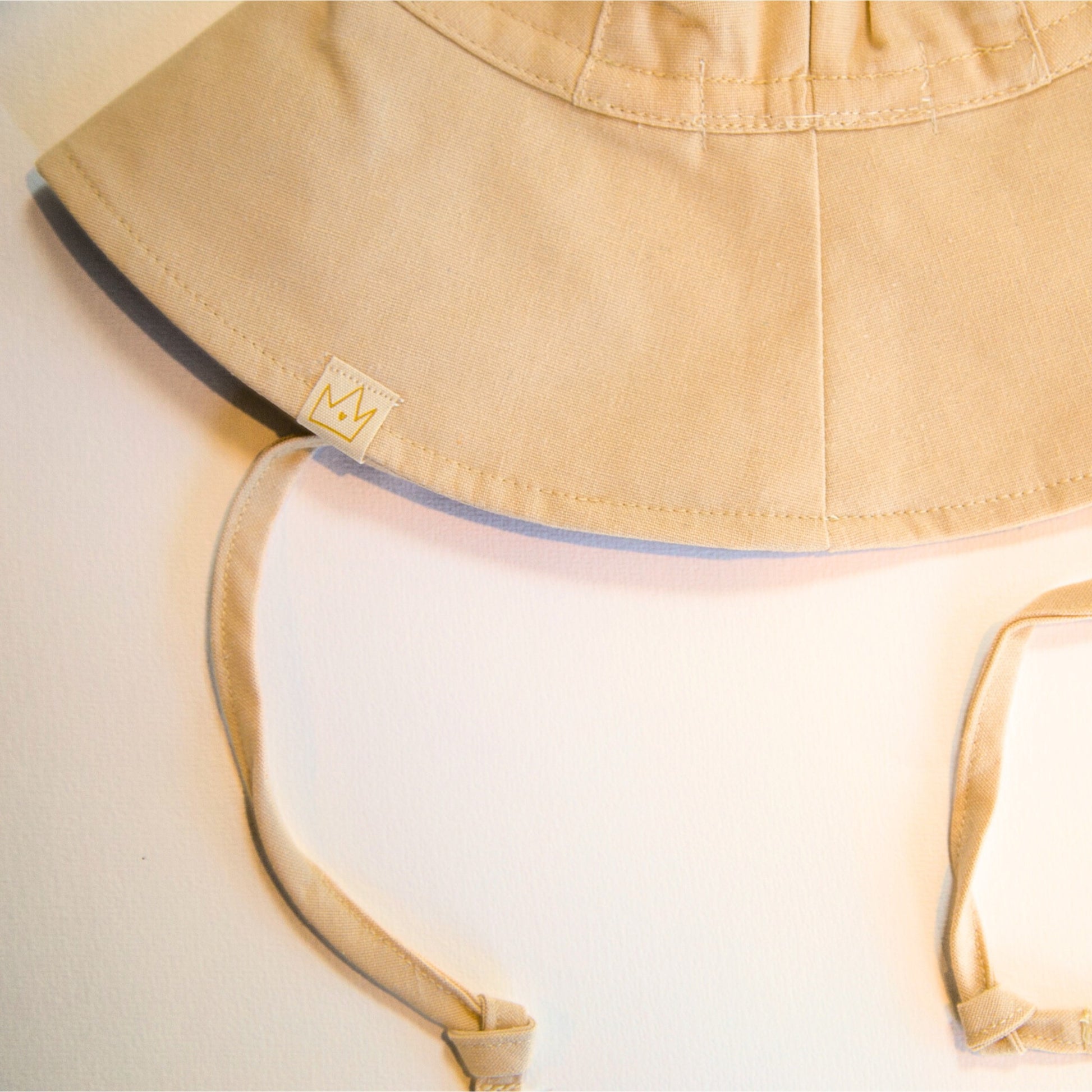 Back view of Sand Dune beige baby hat showing the logo tag and adjustable neck tie for a secure and comfortable fit